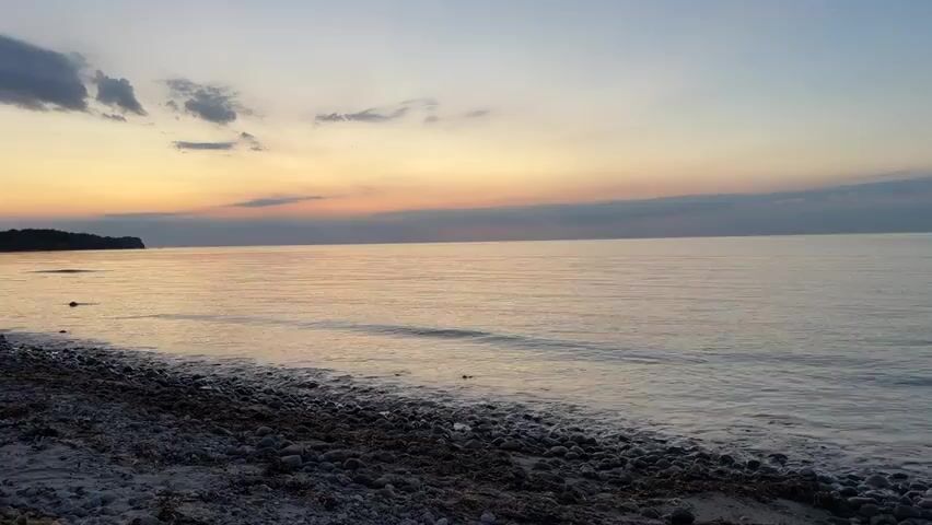 A slow pan across a calm body of water at sunset. The sky displays a gradient of colors from pale yellow near the horizon to a soft blue at the top. A thin band of orange and pink hues are visible just above the horizon. The water is still, with gentle ripples near the shore. The foreground consists of a pebble beach with scattered seaweed. The overall atmosphere is serene and peaceful.