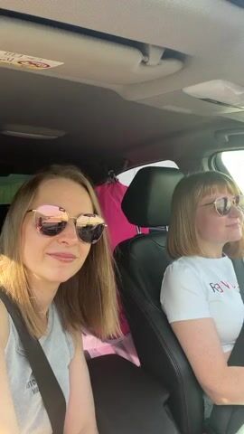 The video shows two women in the front seats of a car. The woman in the passenger seat has short blonde hair and sunglasses, and is wearing a white shirt. The woman in the driver's seat has long blonde hair, sunglasses, and a striped tank top. She is smiling, and runs her fingers through her hair. A pink bag is hanging on the back of the passenger seat. The interior of the car is visible, including the headrests, sun visor, and various stickers. The car appears to be in motion. Music plays in the background.