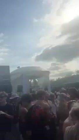 Here is a concise description of the video:

The video shows a crowd of people gathered outdoors, likely at an event or party.  Many of them are facing towards a white structure that appears to be a stage or DJ booth. The day is sunny but cloudy, and the sky is visible above the crowd and the stage. Green trees are visible behind the stage.  The scene is energetic and festive.
