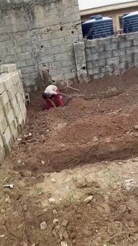 The video shows a man digging a trench in the ground with a shovel. He is wearing red pants and no shirt. The ground is brown dirt with some rocks. There is a brick wall in the background, and a building with water tanks on top. Another person in a yellow shirt is also digging in the distance. The scene appears to be an outdoor construction or excavation site.