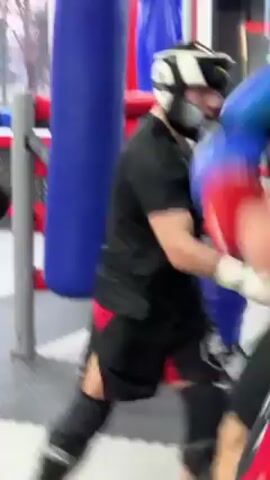 Two men are sparring in a boxing ring. One is wearing red gloves and the other is wearing blue gloves. They are both wearing black shirts. The man with red gloves is punching the man with blue gloves. The man with blue gloves is blocking the punches. The ring is surrounded by red and white striped poles. There is a blue punching bag in the background.