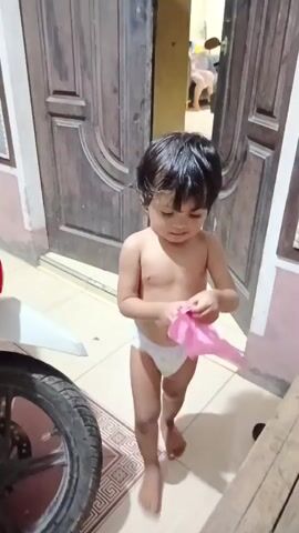 A young child, likely a toddler, stands shirtless and wearing a diaper. The child has dark hair and is holding a crinkled piece of pink paper, seemingly playing with it. They appear to be indoors, with wooden doors and tiled flooring visible in the background. To the left, part of a motorcycle tire is visible, suggesting the setting might be a home or a yard. The lighting appears to be natural, possibly indicating daytime. The child's expression is one of curiosity or concentration. There are no discernible timestamps or readable text within the frame. The overall scene conveys a sense of casual, everyday life.
