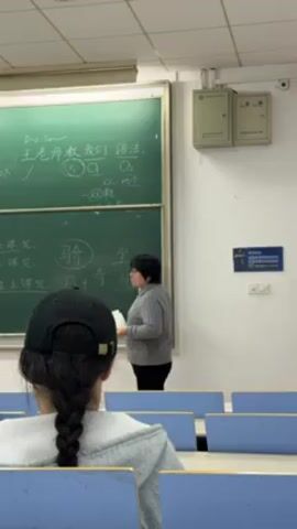 The video shows a classroom setting with a teacher at the chalkboard and a student sitting in the foreground with their back to the camera. The teacher, an Asian woman with short dark hair, is wearing a gray sweater and glasses. She is writing on a green chalkboard covered in Chinese characters and diagrams, referencing a paper in her left hand. The student in the foreground is wearing a gray hooded sweatshirt and a black baseball cap, and their hair is braided down the back. The classroom has rows of blue chairs and fluorescent lighting. There are speakers mounted on the wall, and a notice board on the right side of the frame. The scene appears to be a language lesson focused on Chinese characters and grammar.
