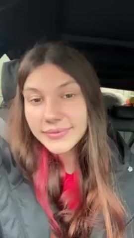 A young woman with long, brown hair sits in a car. She is wearing a black puffer jacket over a red shirt. She has a nose piercing and her hair is dyed pink at the ends. She looks directly at the camera and speaks. The interior of the car is visible in the background.