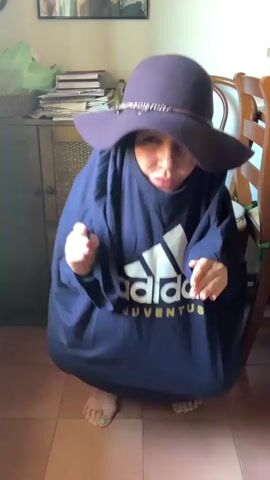 This media file features a person dressed in an oversized navy blue Adidas Juventus t-shirt and a wide-brimmed purple hat, crouching on a tiled floor. The person's face is partially obscured by the hat, but their mouth is pursed and their hands are held up in a playful gesture. The t-shirt is so large that it envelops their entire body, creating a rounded silhouette. The Adidas logo is prominently displayed on the front of the t-shirt in white and gold. The scene appears to be indoors, with a wooden cabinet and stacks of papers visible in the background. A wooden chair is also visible to the right. The lighting suggests it could be daytime. The person's posture and facial expression convey a sense of playfulness and perhaps a touch of humor. The visible text includes "Adidas" and "Juventus." The location context indicates this scene is in San Quirico d'Orcia, Italy.