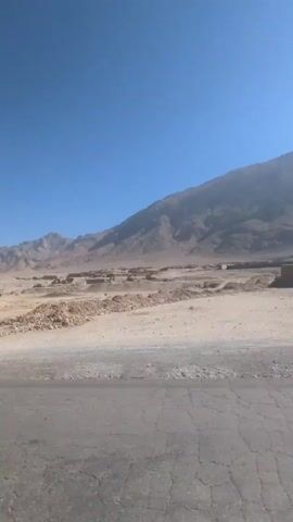 The video shows a drive through a desert landscape with mountains in the background under a clear blue sky. The foreground includes a paved road and a barren, rocky desert terrain. In the distance, there appear to be some small, rectangular structures, possibly buildings, scattered across the landscape. The overall impression is one of a dry, remote, and sparsely populated area.