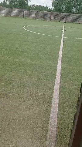 The camera pans across a fenced green soccer field. A boy in black clothing walks across the soccer field, away from the camera. Behind the fence there is a grey brick building, telephone poles, and trees. The sky is overcast.