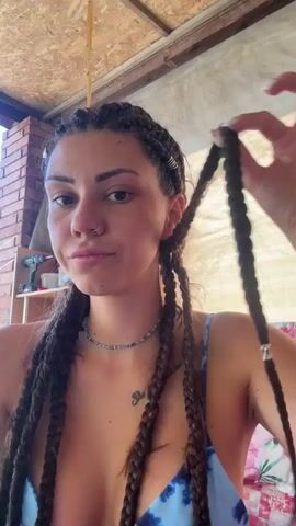 A young woman with dark braided hair is shown in a medium close-up shot, holding a pair of blue-handled scissors. She is in the process of cutting one of her braids, holding the scissors near the end of the hair strand. Her expression is neutral, perhaps a bit mischievous, as she looks towards the camera. She is wearing a blue and white tie-dye bikini top and a beaded necklace. A tattoo is visible on her chest and another on her left wrist.

The background suggests an outdoor or semi-outdoor setting. There is a wooden ceiling or roof structure above, and a brick wall on the left. A glimpse of a white tarp or fabric is visible behind her. The lighting appears to be natural, suggesting it is daytime, possibly sunny. The overall scene has a casual, relaxed feel. There is no visible text in the media. The activity is clearly the woman cutting her own hair, specifically her braids. The focus is on the act of cutting and the woman's action.