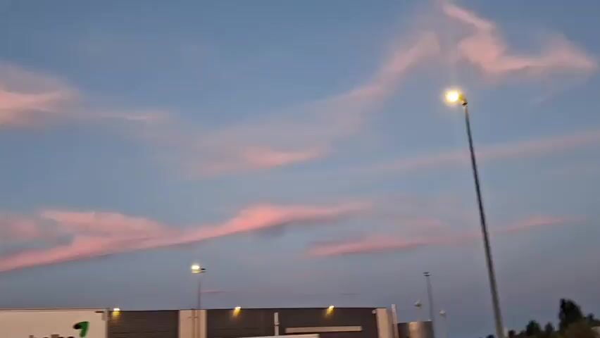 The video shows a sky with pink and orange clouds at sunset. In the foreground, there are several parked trucks. The camera pans from left to right, revealing more of the trucks and the surrounding landscape, which appears to be a rest stop or truck depot. There are streetlights visible, illuminating the scene. The overall impression is of a peaceful evening at a transportation hub.