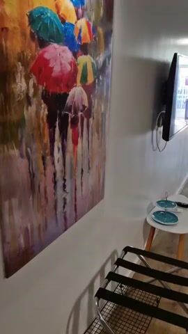 The video pans across a hallway and into a living/dining area. On the left wall of the hallway is a large painting of colorful umbrellas. A television is mounted on the wall above a small white side table with two blue plates and a remote. A luggage rack is in the foreground. The hallway leads to an open doorway showing a room with a blue chair and a picture on the wall. The video then pans right into the living/dining area. A sofa with colorful pillows is against the gray wall, with a painting of colorful parrots above it. A round dining table with a tablecloth and two yellow chairs is in the center of the room. On the table is a vase with yellow flowers and some papers. To the right of the table is a red console table with a mirror above it. A small cabinet is partially visible on the far right. The room has wooden floors and gray walls.