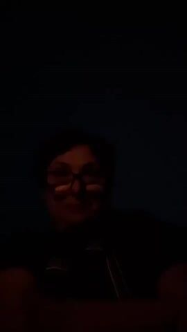 A person wearing glasses and a dark shirt is shown. The background is dark, suggesting low lighting. The person appears to be looking directly at the camera, with a slight smile.