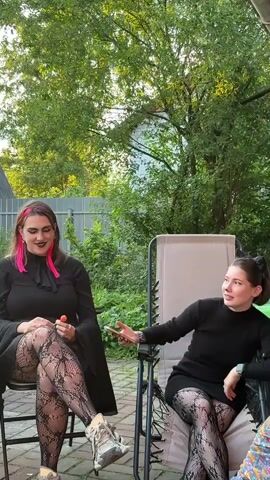 Two women are seated in chairs outdoors. The woman on the left is wearing a black dress, fishnet stockings, and sneakers. She has bright pink hair extensions and is holding a lollipop. The woman on the right is wearing a black dress, fishnet stockings, and has cat ears on her head. She is holding a phone in her left hand and gesturing with her right hand. They are in a yard with trees and foliage in the background. The woman on the right appears to be speaking.