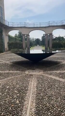 This media file depicts a tranquil courtyard scene in Varese, Italy. The focal point is a large, dark, conical water feature situated in the center of a cobblestone patio. The cobblestones are arranged in a radiating pattern, with a narrow drainage channel running from the water feature towards the viewer.

Behind the water feature, a grand architectural structure with three arched openings forms a backdrop. Each arch reveals a glimpse of a formal garden path leading towards a distant fountain. The arches are supported by tall, classical columns, and a decorative wrought-iron railing runs along the top of the structure. Lush green hedges and mature trees frame the view through the arches, suggesting a well-maintained garden estate.

The time of day appears to be daytime, with soft sunlight casting shadows from the water feature onto the cobblestones, creating a starburst effect. The sky is a pale blue with a few scattered clouds, indicating pleasant weather. The overall atmosphere is serene and elegant, characteristic of Italian gardens and architecture. There are no people visible in the frame, emphasizing the stillness of the location. The scene evokes a sense of peace and historical grandeur. No visible text is present.