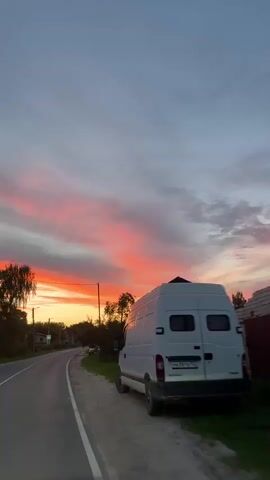 A white cargo van is parked on the side of a rural road, facing away from the camera. The road curves to the left, disappearing into the distance. The sky is a vibrant mix of pink, orange, and gray clouds, suggesting a sunset or sunrise. Trees and houses line the road, with some visible streetlights and traffic signs. The scene appears to be in a rural or suburban area, possibly in Eastern Europe, given the style of the houses and the van. The lighting and atmosphere suggest a peaceful, possibly contemplative moment.
