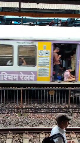 The video shows a train with open doors at a station platform. Several people are standing in the doorways and inside the train cars, with some appearing to be boarding or disembarking. The train is white and purple, with text on the side that appears to be in a non-English script. A metal fence separates the platform from the train tracks. In the foreground, a person wearing a white shirt and a cap is walking away from the camera. The platform is made of concrete and gravel. The overall scene depicts a busy train station environment.