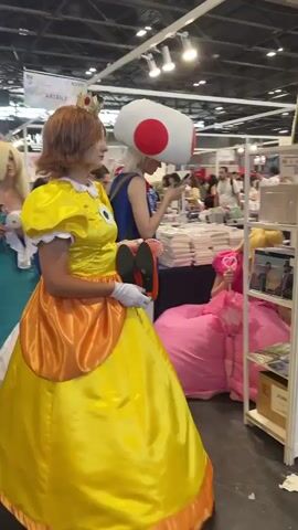 The video shows a crowded indoor event, likely a convention or exhibition. In the foreground, a person in a yellow Princess Daisy costume from the Super Mario franchise is visible, turning their head to the right. Behind them, a person wearing a large Toad head costume from Super Mario is also visible. The environment appears to be a large hall with booths and displays. In the background, other attendees can be seen walking around, some dressed in cosplay. The camera is panning and moving, indicating that the video is likely captured by someone walking through the event. The lighting is bright, typical of an indoor venue. The overall impression is a lively and busy public gathering focused on pop culture.