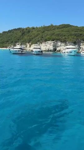 This video captures a picturesque scene of a turquoise bay on a sunny day in Ozias, Greece. The foreground is dominated by the crystal-clear, shimmering blue water, revealing shadows and the seabed beneath. In the mid-ground, several white motorboats are anchored close to a white rocky cliff face and a lush green hillside. Further in the distance, a white sailboat with its mast prominently visible is sailing across the bay. Beyond the boats, a sandy beach is visible, dotted with people and what appear to be small structures or umbrellas, suggesting a populated tourist destination. The background is a verdant, tree-covered hillside rising steeply from the coast under a clear, bright blue sky. The overall impression is one of a serene and idyllic summer day at a popular coastal resort, with opportunities for swimming, boating, and beach activities. The time of day is clearly daytime, likely mid-morning to early afternoon, given the bright sunlight and clear skies. There is no visible text in the video.