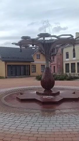 A fountain is seen with water spraying from the top. The fountain has a reddish-brown base with a teardrop-shaped element in the center. The fountain is surrounded by a circular basin and a brick-paved area. In the background, there are buildings with tiled roofs and windows. The sky is overcast.