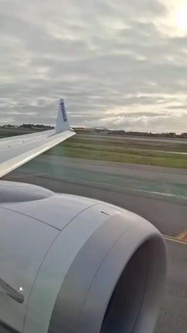The view from an airplane window shows the wing and engine of a Ryanair plane as it speeds down a runway. The wing is white with the "RYANAIR" logo visible on the winglet. The engine is gray and cylindrical, with a dark center. The runway is gray asphalt, and beyond it is a grassy area with some standing water. In the distance, a bridge-like structure can be seen. The sky is overcast with a mix of gray and white clouds, and a hint of sunlight peeks through.