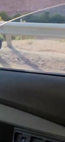 The video shows a view from inside a vehicle, looking out a window. The scene outside is a roadside with a guardrail and vegetation. The road surface is visible, with a yellow line marking the lane. Shadows are cast on the road, indicating sunlight. The environment appears to be a rural or undeveloped area, possibly mountainous, with dry vegetation and rocky terrain in the background. The footage is slightly blurred, suggesting movement. There are no people or specific objects of interest clearly discernible within the frame.