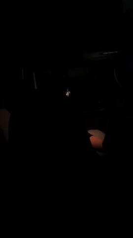 The video displays a dark room with minimal illumination. In the center, a person's face is briefly illuminated by a faint light source, possibly a phone screen. Their eyes are visible, looking towards the viewer. To the right, another person is partially visible, their hand resting on a glowing red object, possibly a light. The background is indistinct due to the darkness.