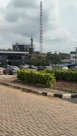 The camera pans across a street scene. The foreground is a paved road with a curb. To the right of the road is a green bush. In the middle ground, there are several cars parked near a modern building with large glass windows and a flat roof. A tall blue sign for "Polaris Bank" is visible. Behind the bank, there is a hill covered with trees. In the background, there is a building under construction with scaffolding, a tall communications tower, and another building with the letters "IFE" on it. A small truck drives by, and a pedestrian walks along the road. The sky is cloudy and overcast.