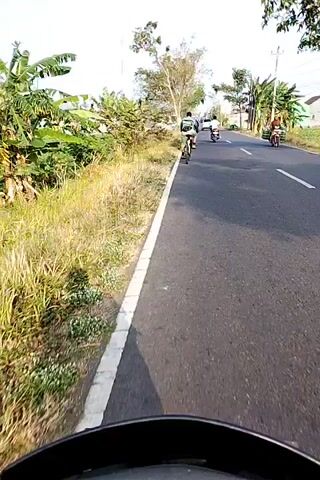 The video begins with a first-person perspective of someone riding a motorbike on a paved road. To the left of the road, there is a grassy and vegetated area. In front of the motorbike, a cyclist is moving away from the camera along the right side of the road. Further ahead on the road, another person is riding a motorcycle. The scene appears to be in a rural or semi-rural area with trees and vegetation lining the road. The sky is bright, suggesting it is daytime, possibly morning or late afternoon due to the angle of the sunlight. The video captures the movement of these vehicles on the road and the surrounding environment.