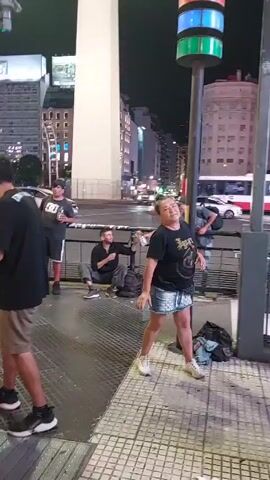 The video shows a public urban scene at night, possibly in a city center. Several people are present. A woman wearing a black t-shirt and denim shorts is seen dancing and gesturing. Three other people are nearby. One is standing, holding a beverage, another is kneeling and appears to be organizing items from a bag, and the third is standing and looking around. Bags are lying on the ground near them. In the background, there's a view of a busy street with cars and buses. The environment includes a tall obelisk-like structure, buildings, street lights, and a traffic signal. The ground is paved.