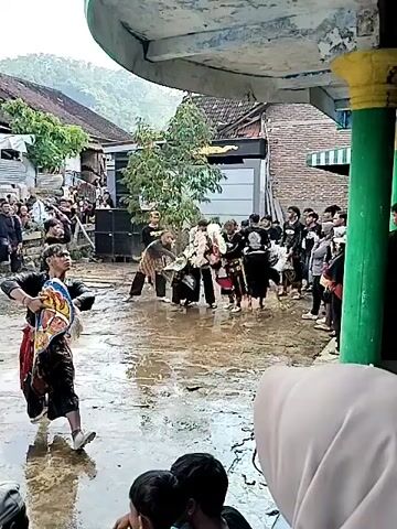 The video shows a traditional Indonesian dance performance taking place in a flooded area. A crowd of people, mainly young men, is gathered around the performers. Some of the performers are wearing traditional black costumes and holding brightly colored props. One performer is lying on the ground, while others are standing or moving around him. The area is flooded with water, and there are houses and trees in the background. The video is shot from under a roofed structure, with pillars visible on the right side of the frame. The weather appears to be overcast or rainy.