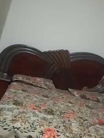 THE VIDEO SHOWS A CLOSE-UP VIEW OF A BED WITH A FLORAL PRINT COMFORTER. TO THE RIGHT OF THE BED IS A DRESSER WITH A LARGE OVAL MIRROR. BEYOND THE DRESSER IS A DOORWAY LEADING TO ANOTHER ROOM. THE ROOM APPEARS TO BE A BEDROOM. THE LIGHT IS DIM.