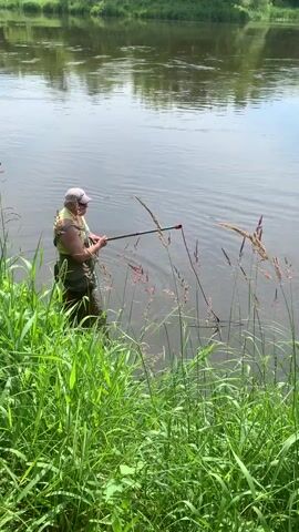 A person stands in shallow water near the edge of a river. They are wearing waders, a vest, and a hat. They hold a long pole with a net attached to the end, which they are using to scoop something from the water. The water is calm and reflective, with green vegetation on the opposite bank. The person appears to be fishing or collecting something from the riverbed.