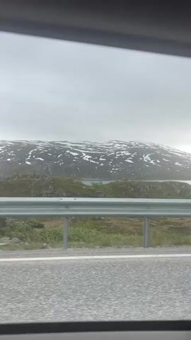 The video shows a view from a vehicle moving along a paved road. The camera is pointed towards a landscape featuring a body of water and a snow-capped mountain range under a cloudy sky. A metal guardrail separates the road from the embankment leading to the water. The terrain is rocky and sparsely vegetated with patches of green and brown.