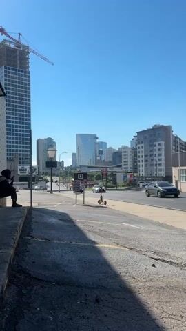 A vertical video shows a street scene with a cityscape in the background. On the left, a person sits on a concrete ledge, looking at their phone. A construction crane is visible above a tall building. The street has cars and an electric scooter. Several buildings of varying heights make up the cityscape, with a large, modern glass building in the center. The sky is clear and blue.
