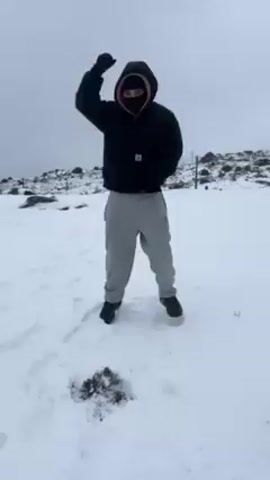 Here is a description of the video:

A person stands in a snowy field wearing a black hooded jacket, a black face covering, and grey sweatpants. They pump their fist in the air and then cross their arms. The background is an overcast sky and a rocky, snow-covered landscape. Music plays throughout the video. There are footprints in the snow.