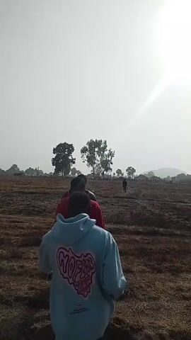 Two people are walking away from the camera across a dry, grassy field on a sunny day. The person closest to the camera is wearing a light blue hoodie with a pink and black design on the back. The second person is wearing a red jacket. Several other people are visible in the distance, scattered across the field. There are a few trees in the middle distance and a hazy horizon. The sky is overcast and the sun is bright, causing lens flare.
