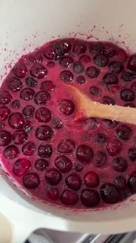 A close-up video shows a white pot filled with dark red berries that appear to be cooking. A wooden spoon is stirring the berries, which are bubbling and releasing a pink juice. The pot is on a stovetop, with the edge of a burner visible at the bottom of the frame. The berries are round and plump, and some of them appear to be bursting as they cook. The liquid is a vibrant pink color, and there are small bubbles on the surface. The spoon is moving in a circular motion, and it is coated in the berry juice.