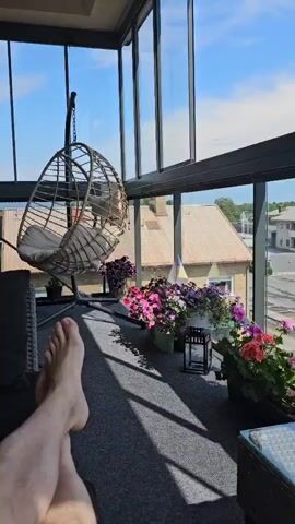 The video shows a view from inside a sunroom looking out at a town. A person's feet and legs are visible in the foreground, resting near a hanging chair. The sunroom has large windows that offer a clear view of the sky and the town outside, which includes buildings, a street, and a parked car. The sunroom is decorated with several potted flowering plants. A glass-topped table with a green candle is also visible.