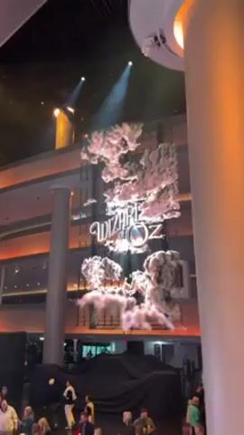 In a high-ceilinged indoor venue, a large animated sign for "The Wizard of Oz" hangs suspended. The sign is framed by clouds and illuminated by spotlights from above. Below the sign, a crowd of people gathers, some standing near a large object covered in black fabric. The environment is modern, with multiple levels visible and cylindrical support columns. The color palette is dominated by beige, white, and black, with orange accent lighting on the ceiling.