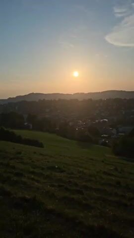 A scenic view of a sunset over a town nestled in a valley. The sun, a bright orange orb, is low on the horizon, casting a warm glow across the sky and silhouetting the distant hills. In the foreground, a grassy hill slopes downwards, its texture detailed by the evening light. The town itself is a collection of buildings and trees, appearing somewhat indistinct in the distance due to the haze and the low sun. The sky is a gradient of blue and orange, with a few wispy clouds.