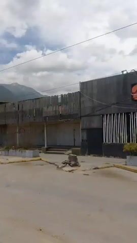 A video shows a street scene with buildings and mountains in the background. The buildings appear to be shops with metal shutters. A few people are visible walking on the street, and a car is parked. The sky is cloudy. The scene suggests an urban or semi-urban environment.