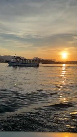 The video captures a serene sunset scene over calm waters, likely in Poros, Greece. The golden sun is low on the horizon, casting a bright, shimmering path of light directly across the water towards the viewer.

In the mid-ground, a white passenger-style motorboat with blue windows is seen moving from left to right, leaving a distinct wake. The camera appears to be on another moving boat, as indicated by the ripples and small waves in the immediate foreground.

The background features a coastline with hills or mountains silhouetted against the bright sunset. Several other boats, including sailboats with visible masts, are scattered across the distant water. The sky above is a mix of warm orange and yellow near the sun, transitioning to softer blues and grays with scattered clouds further up. A small bird can be seen flying high in the sky early in the clip.

The overall atmosphere is peaceful and picturesque, showcasing boating activity during a beautiful sunset. There is no visible text within the video.