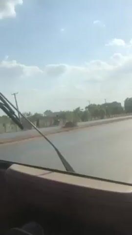 From the perspective of inside a car, the video shows a view of a wide, two-lane road under a partly cloudy sky.  The road is mostly empty, but a three-wheeled vehicle (likely an auto-rickshaw) and a red car pass by. Trees and low buildings line the side of the road behind a low wall. The dashboard of the car is visible in the foreground.