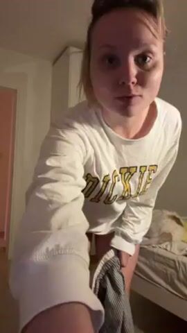 A young woman with blond hair pulled back is standing in a bedroom. She is wearing a white long-sleeved shirt with “DICKIES” printed on the front. She is pulling down striped pants. A bed is visible behind her, along with a white cabinet and a partially visible pink door. She is facing the camera with a neutral expression.