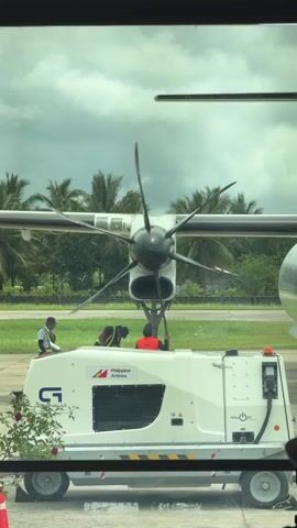From an indoor vantage point looking through a window, the video shows a Philippine Airlines airplane on the tarmac. The camera focuses on the front of the plane, specifically its large propeller with six blades. Three ground crew workers are visible near the front of the aircraft. One stands to the left of the machine, wearing a light-colored vest over a dark shirt and a hat, possibly observing the other two. The other two workers are kneeling or bending down closer to the front, one wearing a bright orange safety vest. A large white generator unit with the Philippine Airlines logo is parked in front of the plane, obscuring some of the view of the ground crew. The background features a green grassy area with palm trees swaying in the wind. The sky is overcast with grey clouds.