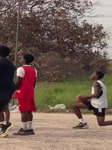 A group of four young men playing basketball on an outdoor court. One of the men, wearing a white tank top and black shorts, kneels on one knee and shoots the basketball towards the hoop. Another man, wearing a red jersey and shorts, stands nearby watching. The other two men are positioned around the court, also observing the shot. The court is surrounded by trees and greenery. The man kneels on one knee to attempt a shot. The ball is a miss.