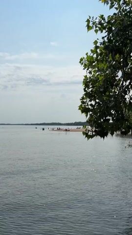 A vertical shot shows a tranquil body of water in the foreground with a sandy spit of land in the middle distance. On the spit, several people are gathered, some wading in the shallow water, others lounging on the sand. A few trees are visible on the far shore, and the sky is partly cloudy. The overall scene depicts a relaxed summer day at a beach.