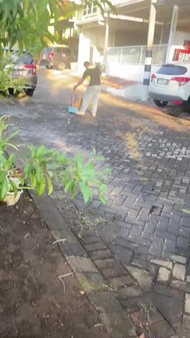 A person is sweeping a paved driveway with a broom. The person is wearing a dark shirt with a yellow stripe and light-colored pants. There are several cars parked along the driveway, and a building is visible in the background. The driveway is made of dark brick pavers. The sun is shining, and there is some greenery in the foreground and background.