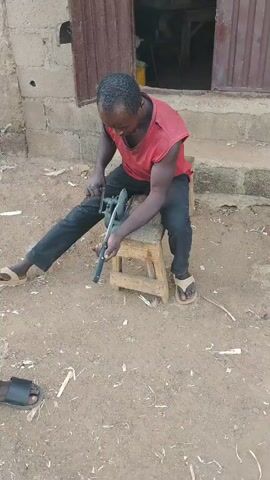 A man in a red tank top and black pants sits on a wooden stool outside a building. He is sharpening a knife with a tool that appears to be a sharpening jig or a vice. The ground is covered in dirt and small pieces of wood. Another person's feet wearing black sandals are visible in the lower left corner of the frame. The building behind the man has a brown door and a brick wall.