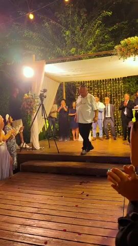 The video captures a wedding ceremony taking place outdoors at night. A man in a light blue short-sleeved shirt and black pants is walking down a wooden aisle, appearing to be walking towards a chuppah. He is smiling and looking down slightly. Several people are visible in the background, including guests applauding on the left and a group of men standing under the chuppah on the right, some of whom are dressed in suits. A videographer is positioned near the chuppah. The chuppah is decorated with white fabric and floral arrangements, and string lights are visible in the background. Red rose petals are scattered on the wooden aisle in front of the man.