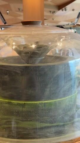 The video shows a close-up of a large, clear glass container with a dark gray base and a transparent lid. Inside the container, a conical object made of dark gray material is suspended, appearing to be part of a grinding mechanism. There is green powder, likely matcha, accumulating on the sides of the container, suggesting the machine is in operation. The powder is spread in horizontal lines, indicating movement and distribution. The background is blurred but appears to be an indoor setting with wooden architectural elements and possibly people in the distance. The overall impression is of a process being observed, possibly a demonstration of a tea grinding machine.